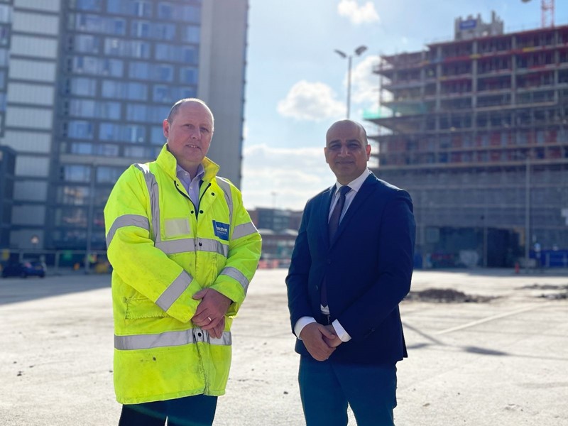 Two men stood on construction site, one in high vis, one in a suit. 