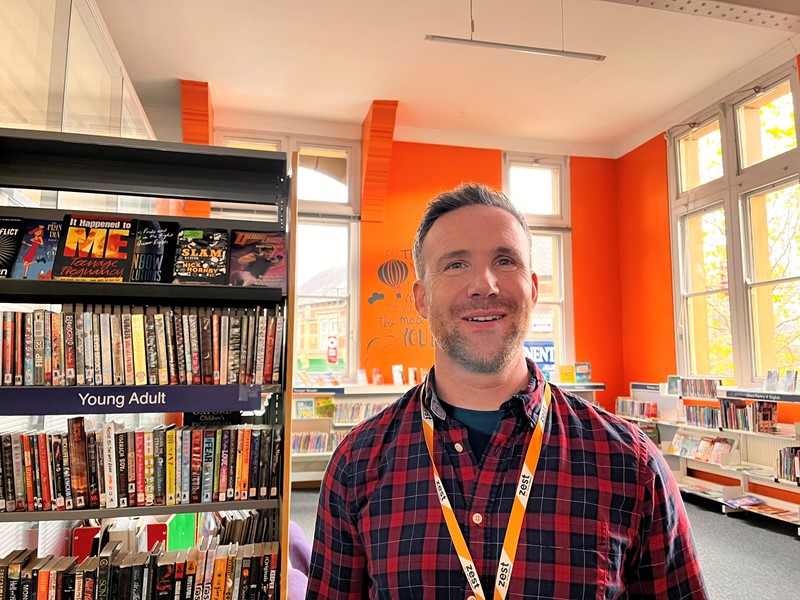 Matt in the library and reception space at The Zest Centre, with orange walls, light coming through windows and shelves with books on 