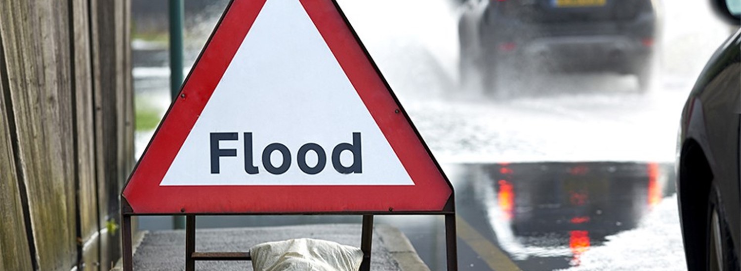 Image of a flood warning sign, in a red triangle on a road where cars are driving through water.