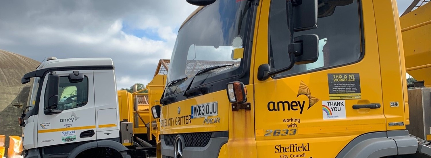 Yellow gritting vehicle in front of white vehicle and cloudy sky
